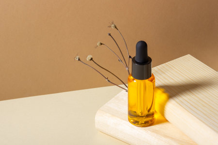 A glass cosmetic bottle with a dropper stands next to a dried flower on a beige background in bright sunlight, on a wooden podium. The concept of natural cosmetics, natural essential oil.の写真素材