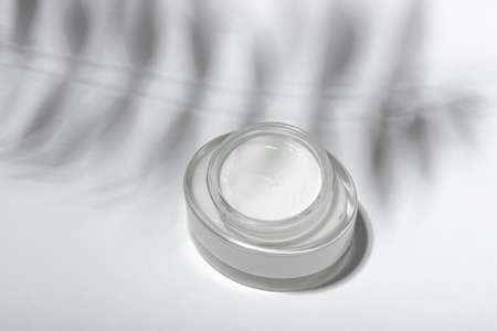 Cosmetic cream or mask in a glass jar with hard shadows from the leaves on a white background. Natural organic care for face and body skin. View from above.の写真素材