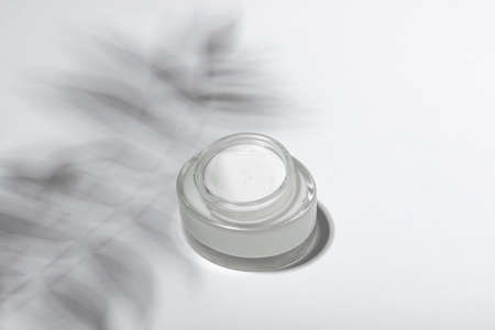 Cosmetic cream or mask in a glass jar with hard shadows from the leaves on a white background. Natural organic care for face and body skin. View from above.の写真素材