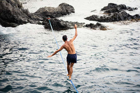 Young dark-haired man walking a tightrope between two rocks over sea water on a sunny day.の写真素材