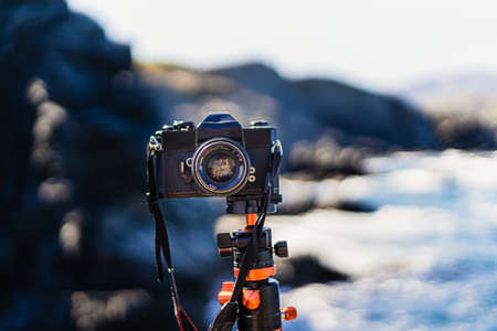 Black unbranded reflex (analog) camera planted on a tripod facing the camera. With a seascape in the background on a sunny summer day.の写真素材