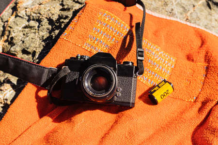 Old unrecognizable black analog photo camera on an orange background with a 35mm reel film next to it. On top of some sea rocks on a sunny day in Spain.の写真素材