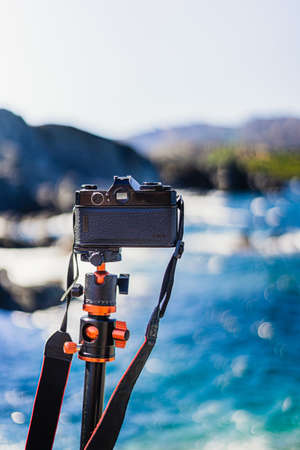 Black (analog) unbranded reflex camera planted on a tripod, taking pictures of the seascape on a...