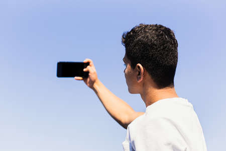 Unrecognizable young brunette man of Maghreb origin, taking a selfie on the beach with his smartphone. Sunny and well-lit summer day, light and blue colors.の写真素材