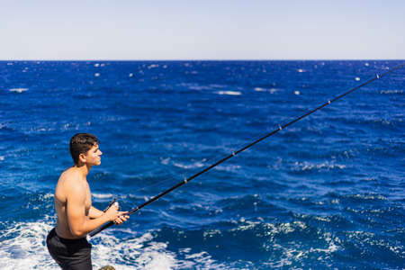 Young dark-haired man of Maghreb origin fishing on his back with the fishing rod raised, on a sunny day and the very blue sky on some sea rocks.の写真素材