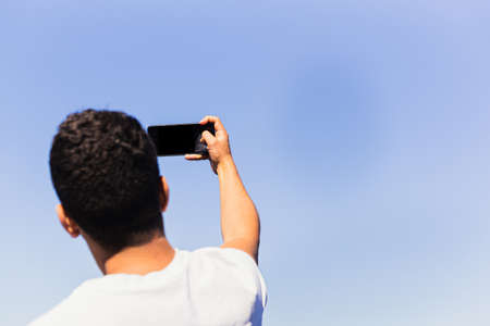 Unrecognizable young brunette man of Maghreb origin, taking a selfie on the beach with his smartphone. Sunny and well-lit summer day, light and blue colors.の写真素材