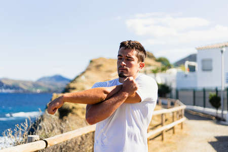 Young sportsman stretching his arms outdoors while looking at the horizon with the sea to the side. Photograph taken from the front on a very sunny day with a Mediterranean landscape.の写真素材