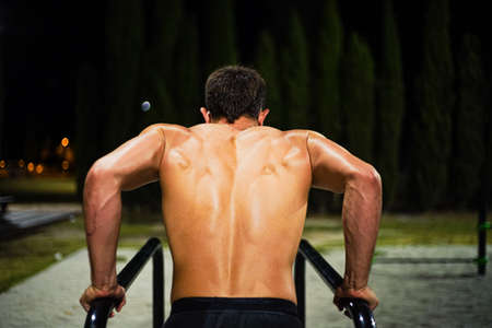 Shirtless muscular young man training at night street workout. In a public bar park. Doing dips....