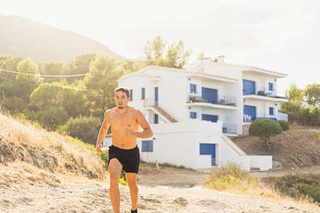 Young sportsman running outdoors on a path. Photograph taken from the front on a very sunny day with a Mediterranean landscape. Model without shirt.の写真素材