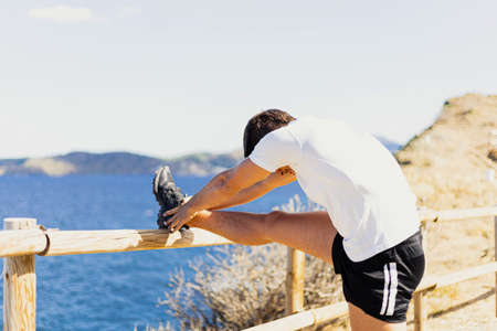 Young sportsman stretching his legs outdoors while looking at the horizon with the sea to the side. Photograph taken from the side on a very sunny day with a Mediterranean landscape.の写真素材