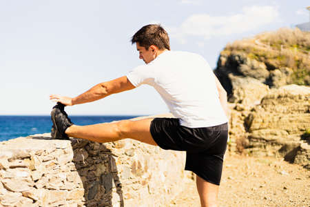 Young sportsman stretching his legs outdoors while looking at the horizon with the sea to the side. Photograph taken from behind on a very sunny day with a Mediterranean landscape.の写真素材