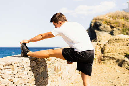 Sportsman stretching his legs outdoors while looking at the horizon with the sea to the side. Photograph taken from behind on a very sunny day with a Mediterranean landscape.の写真素材
