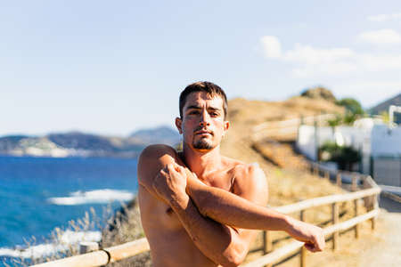Young sportsman stretching his arms outdoors while looking at the horizon with the sea to the side. Photograph taken from the front on a very sunny day with a Mediterranean landscape. Model without shirt.の写真素材