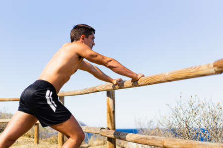 Young sportsman stretching his legs outdoors while looking at the horizon with the sea on the side. Photograph taken from the side on a very sunny day with a Mediterranean landscape. Model without shirt.の写真素材