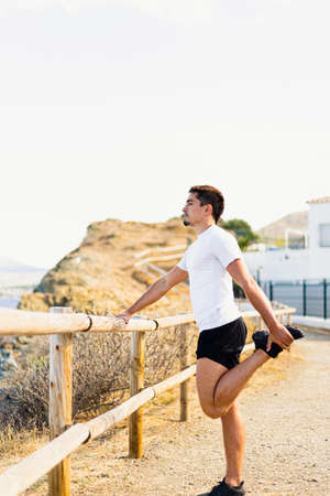 Young sportsman stretching his legs outdoors while looking at the horizon with the sea to the side. Photograph taken from the side on a very sunny day with a Mediterranean landscape.の写真素材
