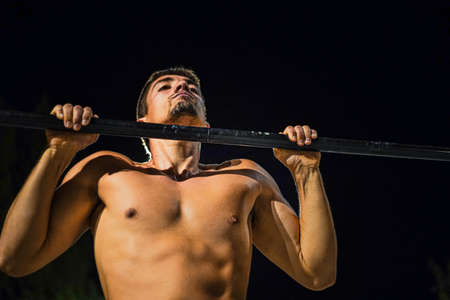 Shirtless muscular young man training at night street workout. In a public bar park. Doing pull-ups....