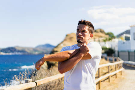 Young sportsman stretching his arms outdoors while looking at the horizon with the sea to the side. Photograph taken from the front on a very sunny day with a Mediterranean landscape.の写真素材