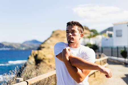 Young sportsman stretching his arms outdoors while looking at the horizon with the sea to the side. Photograph taken from the front on a very sunny day with a Mediterranean landscape.の写真素材