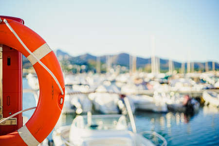 Red lifebuoy photographed up close on a sunny day with a boat harbor and small boats in the...