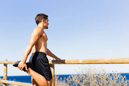 Young sportsman stretching his legs outdoors while looking at the horizon with the sea on the side. Photograph taken from the side on a very sunny day with a Mediterranean landscape. Model without shirt.の写真素材