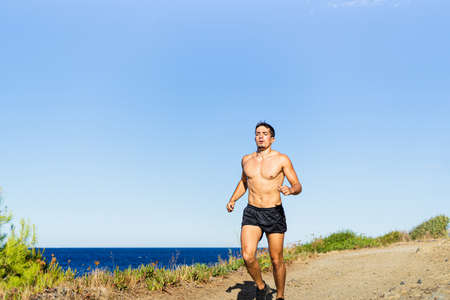 Young sportsman running outdoors on a path. Photograph taken from the front on a very sunny day with a Mediterranean landscape. Model without shirt.の写真素材