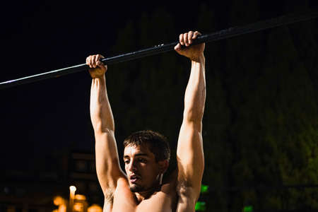 Shirtless muscular young man training at night street workout. In a public bar park. Doing pull-ups. Front photography.の写真素材