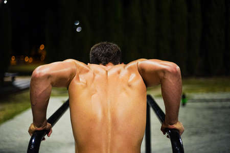Shirtless muscular young man training at night street workout. In a public bar park. Doing dips. Photography from the back.の写真素材