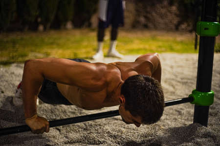 Shirtless muscular young man training at night street workout. In a public bar park. Doing push-ups. Photography from the front.の写真素材