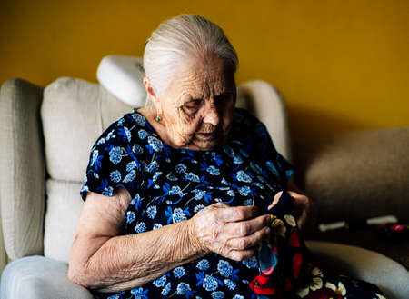 Old grandmother in a blue dress sewing a garment on the sofa at home. Concept of old age and something handmade, handmade.の写真素材