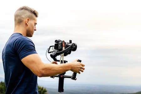 Young muscular Caucasian man manning a camera on top of a gimbal while working recording a scene in the middle of a field. Videomaker with steadicam and professional team working.の写真素材