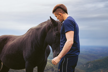 Young Caucasian man with mohawk hair hugging a black horse in the middle of the mountain on a day with cloudy sky.の写真素材