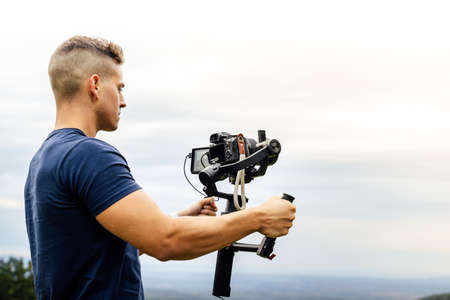 Young muscular Caucasian man manning a camera on top of a gimbal while working recording a scene in the middle of a field. Videomaker with steadicam and professional team working.の写真素材