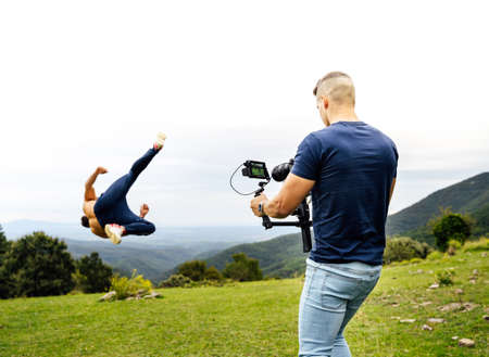 Young and fit cameraman recording with a gimbal a person doing sports, specifically stunts (tricking). Shot from behind, behind the cameras / backstage.の写真素材
