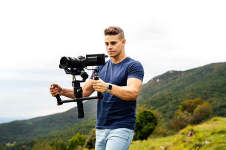 Young muscular Caucasian man manning a camera on top of a gimbal while working recording a scene in the middle of a field. Videomaker with steadicam and professional team working.の写真素材