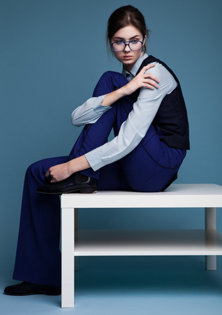 Businesswoman portrait sitting on white table with glasses and blue suit with hand on her shoulder. Shot in studio on blue background.の写真素材