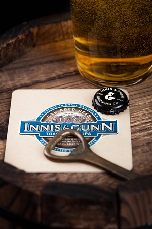 LONDON, UK - AUGUST 10, 2018: Innis & Gunn beer coaster with bottle top and opener and glass of beer on top of wood barrel.