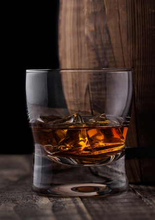 Glass of whiskey with ice cubes next to wooden barrel. Cognac and brandy drinkの写真素材