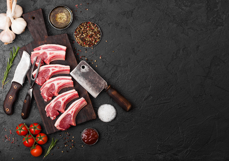 Fresh raw butchers lamb beef cutlets on stone board with vintage meat fork and knife and hatchet on black background.Salt, pepper and oil with tomatoes and garlic and barbecue sauce.の写真素材