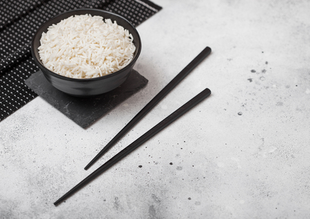 Black bowl with boiled organic basmati jasmine rice with black chopsticks and sweet soy sauce on stone mat on light kitchen table  background.の写真素材