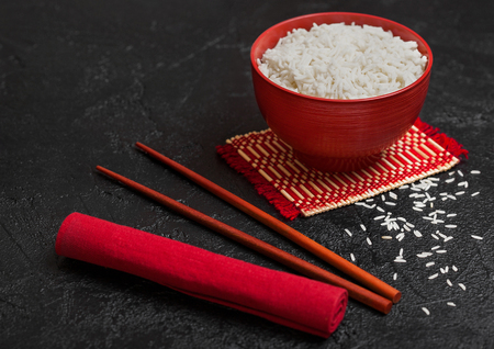 Red bowl with boiled organic basmati jasmine rice with red chopsticks and sweet soy sauce on bamboo placemat with red linen towel on black stone.の写真素材