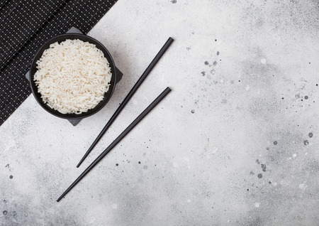 Black bowl with boiled organic basmati jasmine rice with black chopsticks and sweet soy sauce on stone mat on light kitchen table  background.の写真素材