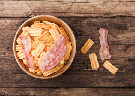 Bacon flavoured snacks chips in wooden bowl with grilled bacon rashers on wood background.の写真素材