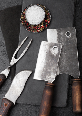 Vintage meat knife and fork and hatchets with stone chopping board and black table background. Butcher utensils.の写真素材