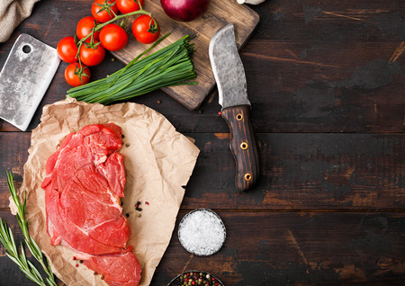 Fresh raw organic slice of braising steak fillet on butchers paper with fork and knife on dark wooden background. Red onion, tomatoes with salt and pepper and herbs.の写真素材