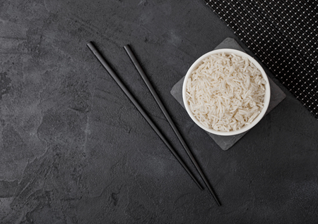 White bowl with boiled organic basmati jasmine rice with black chopsticks on stone mat on black stone background.の写真素材