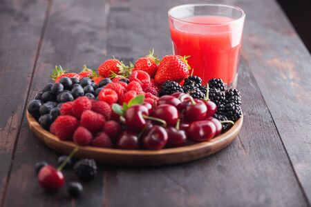 Fresh organic summer berries mix in round wooden tray with glass of juice on light wooden table background. Raspberries, strawberries, blueberries, blackberries and cherries. Top viewの写真素材
