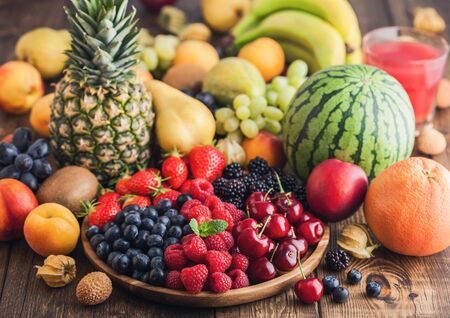 Fresh organic summer berries mix in wooden tray and exotic fruits on wooden background. Raspberries, strawberries, blueberries, blackberries and cherries. Watermelon, pear, pineapple, grapes. Top viewの写真素材