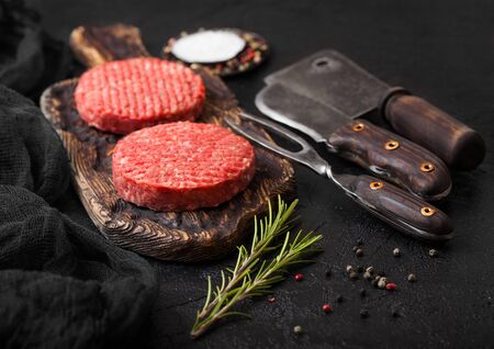 Fresh raw minced homemade farmers grill beef burgers on vintage chopping board with spices and herbs and meat hatchet, fork and knife on black background.の写真素材