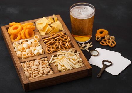 Glass of craft lager beer and opener with box of snacks on dark background. Pretzel,salty potato sticks, peanuts, onion rings with nachos in vintage box with openers and beer mats. Top viewの写真素材