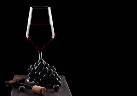Glass of red wine with dark grapes and vintage corkscrew opener and cork on wooden board on black background.の写真素材